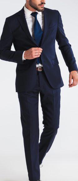 young businessman walking on light grey background and buttoning his navy suit while looking down to side, full body picture